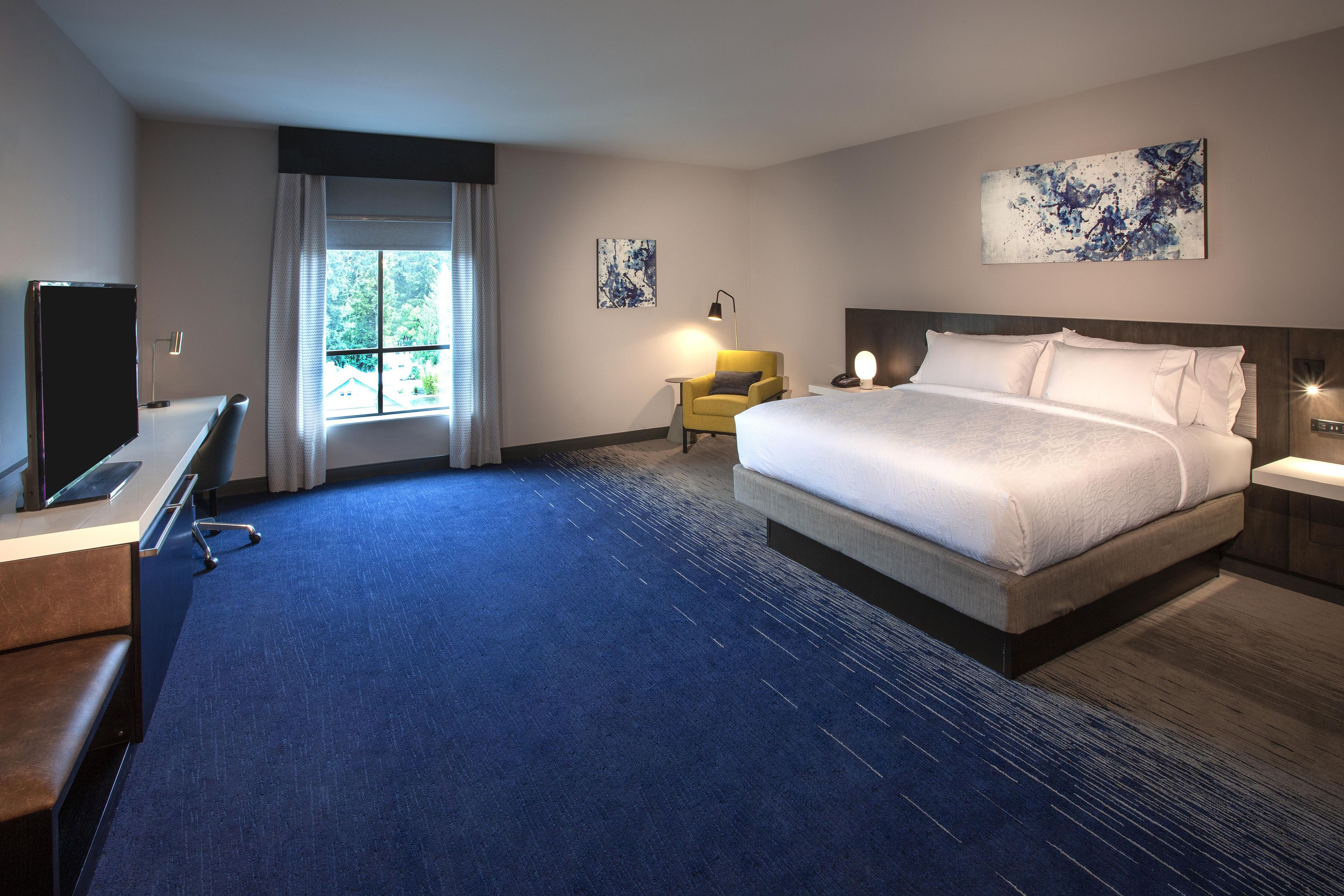A hotel room with a large bed, a desk, a chair, a lamp, and artwork on the walls.