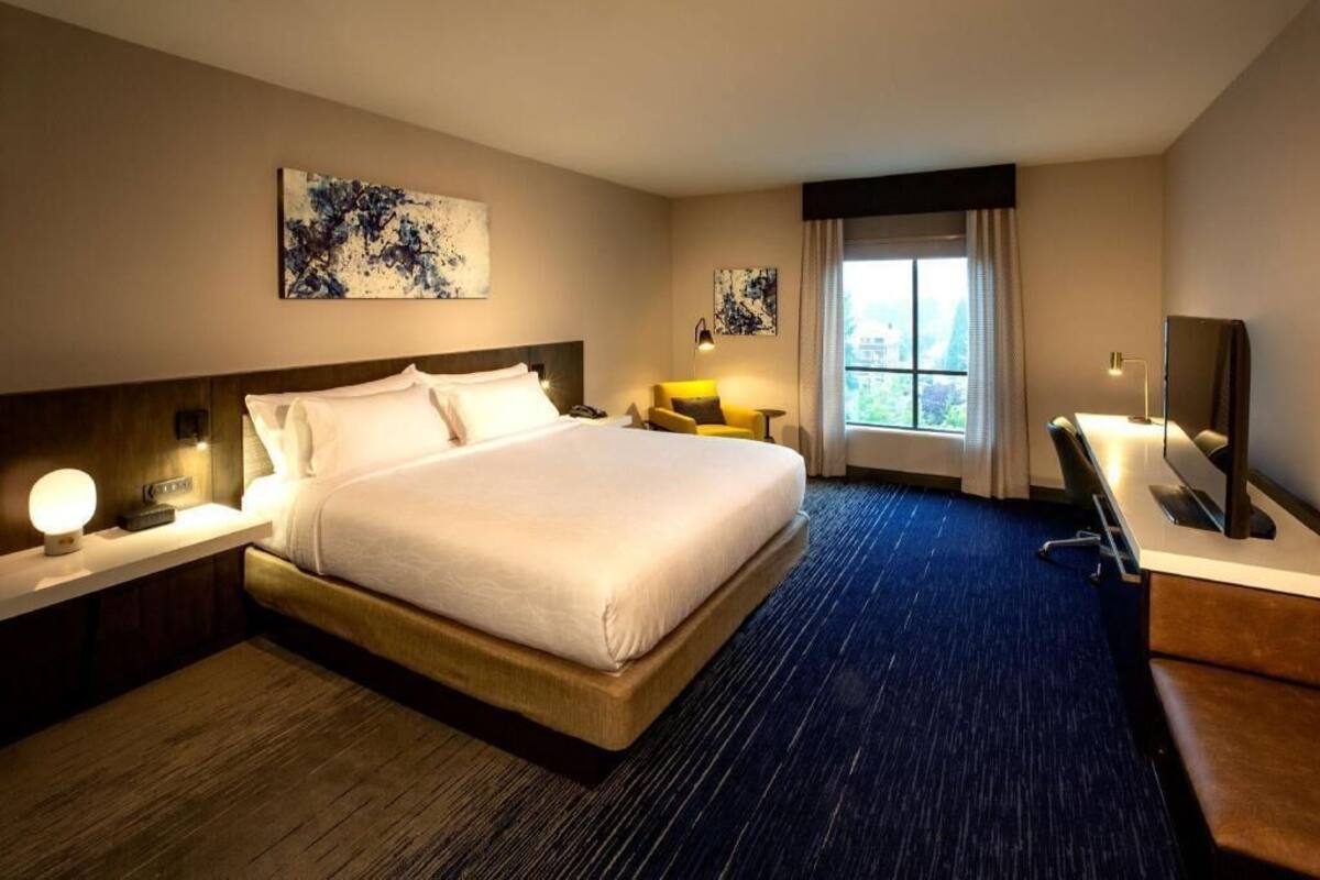 A hotel room with a large bed, bedside tables, a desk, a chair, and artwork on the walls.