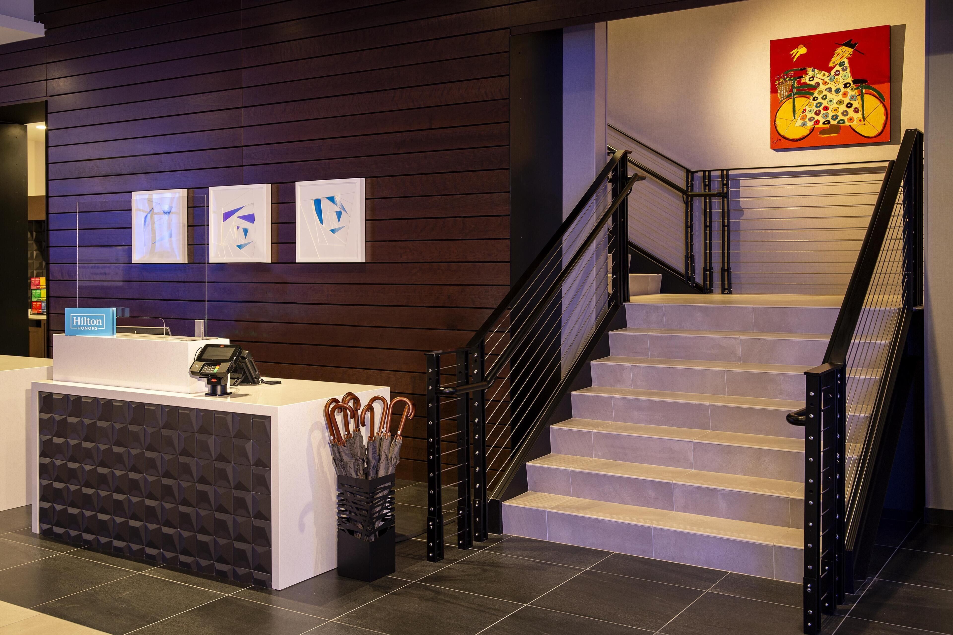 A reception desk with a modern design, a staircase with black railings, and a colorful artwork on the wall.