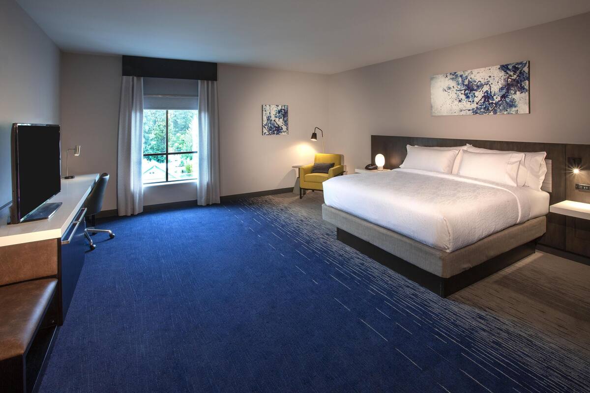 A hotel room with a large bed, a desk with a chair, a small table with a lamp, and artwork on the walls.
