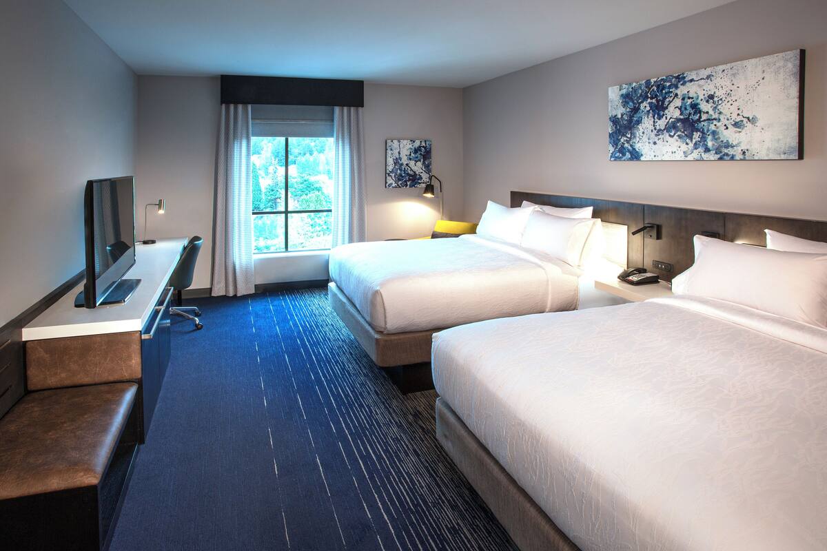 A hotel room with two beds, a desk with a chair, a television, and a large window with curtains.