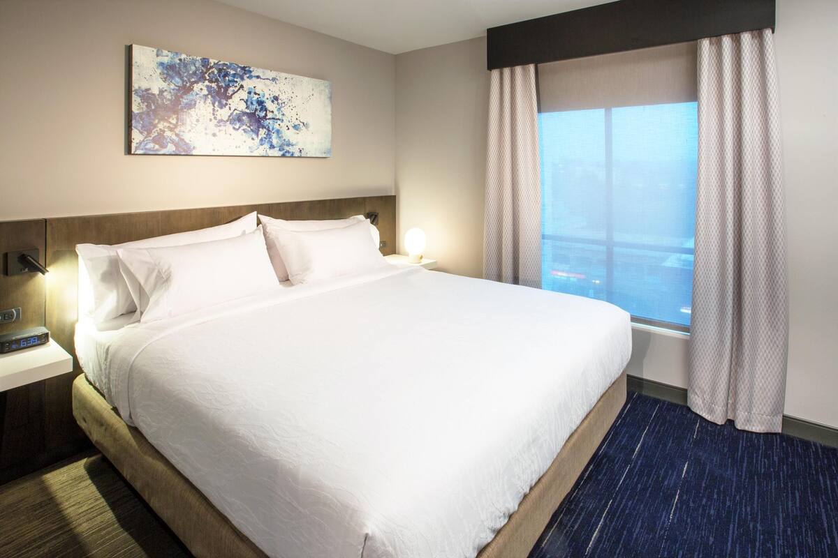 A hotel room with a large bed, a bedside table with a lamp, a window with curtains, and a painting on the wall.