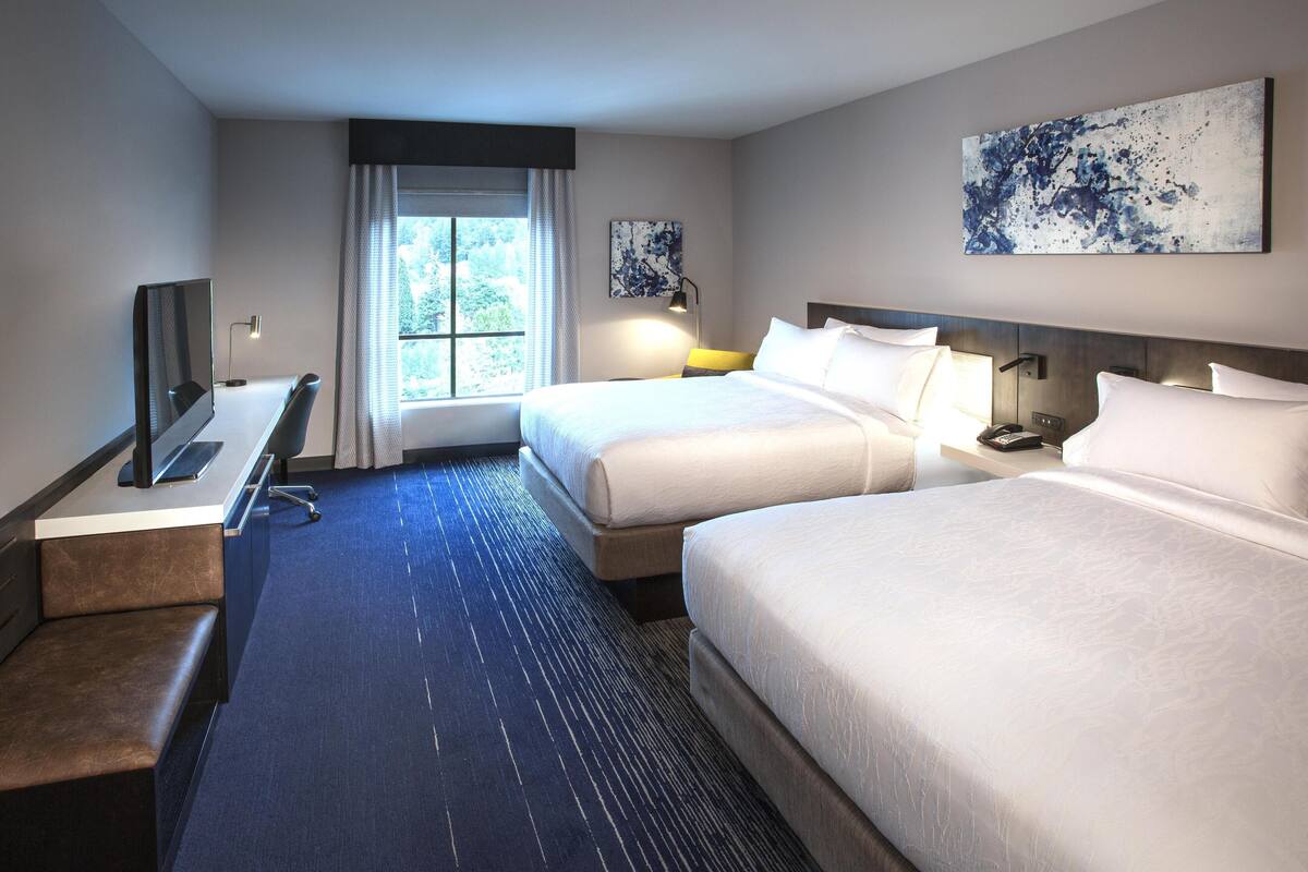 A hotel room with two beds, a desk, a TV, and a window with curtains.