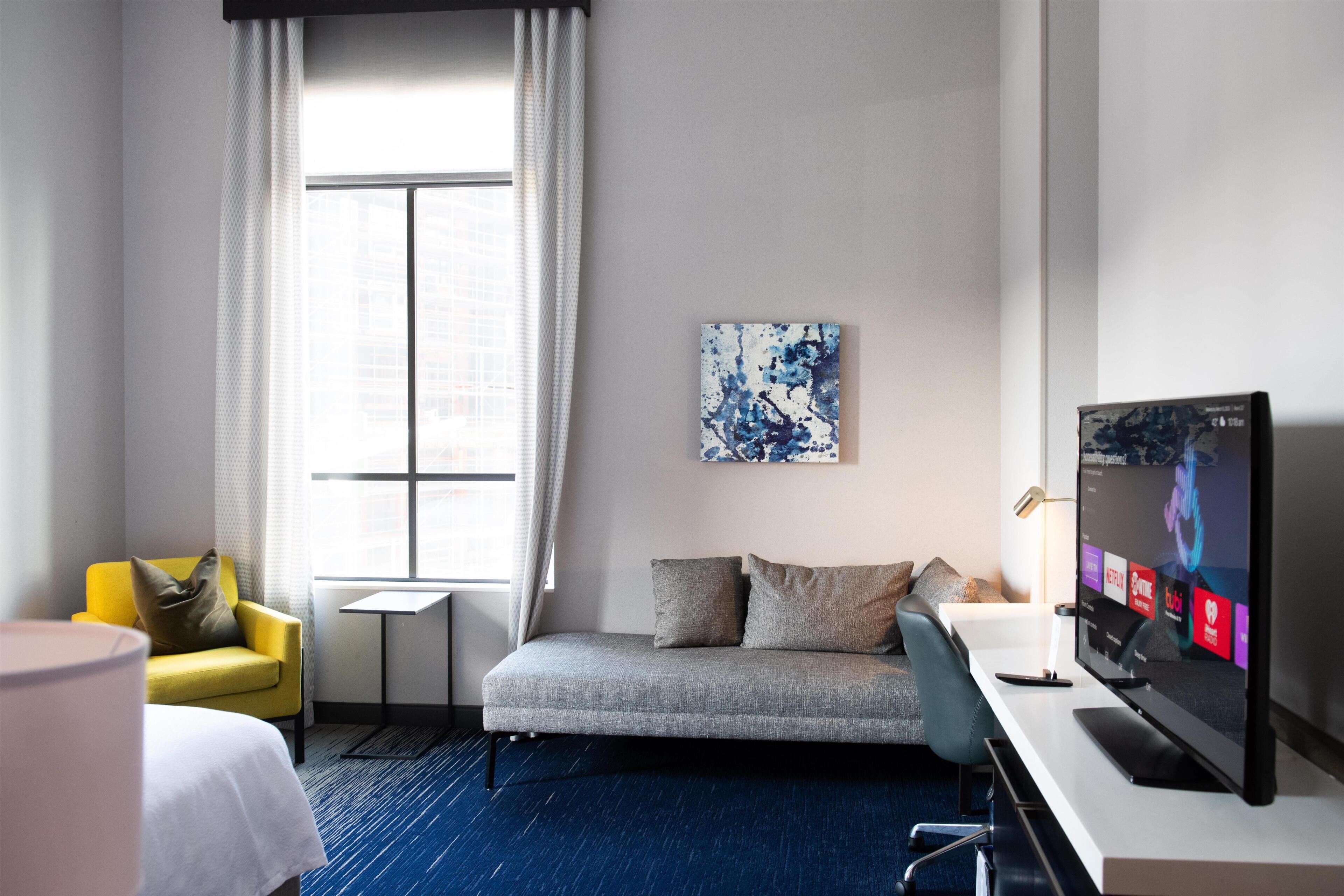 Habitación de hotel con cama, sofá, televisión, una mesita y ventana con cortinas.