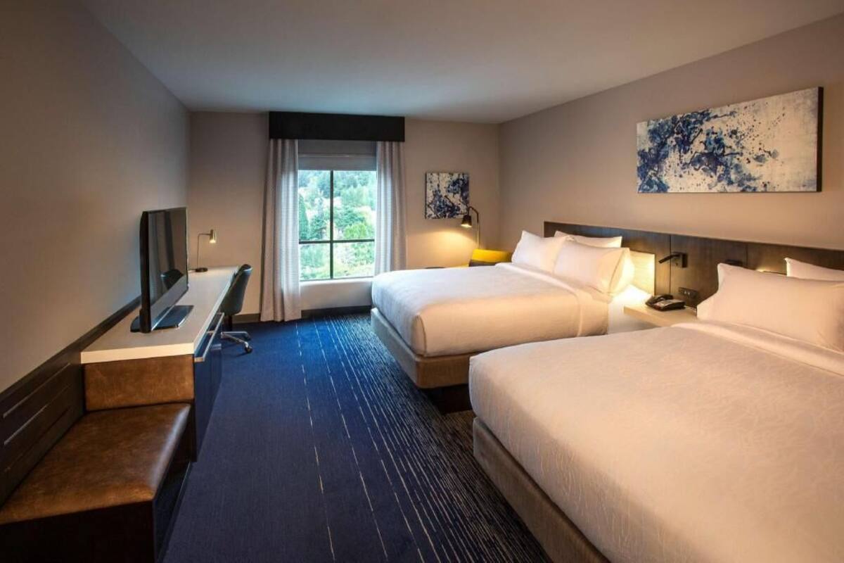 A hotel room with two beds, a desk with a flat-screen TV, a bench, a window with curtains, and artwork on the walls.