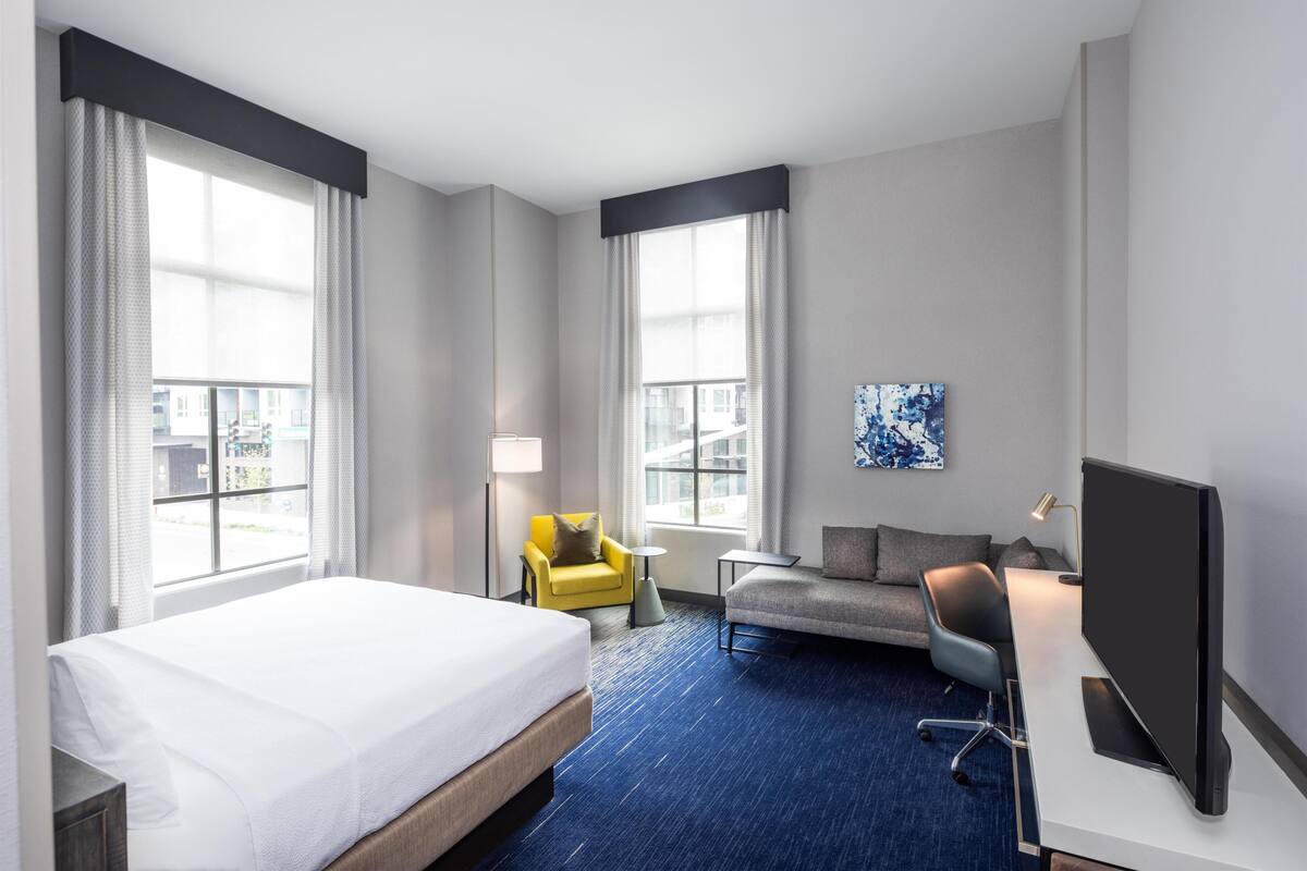 A hotel room with a bed, two armchairs, a desk with a TV, and large windows.