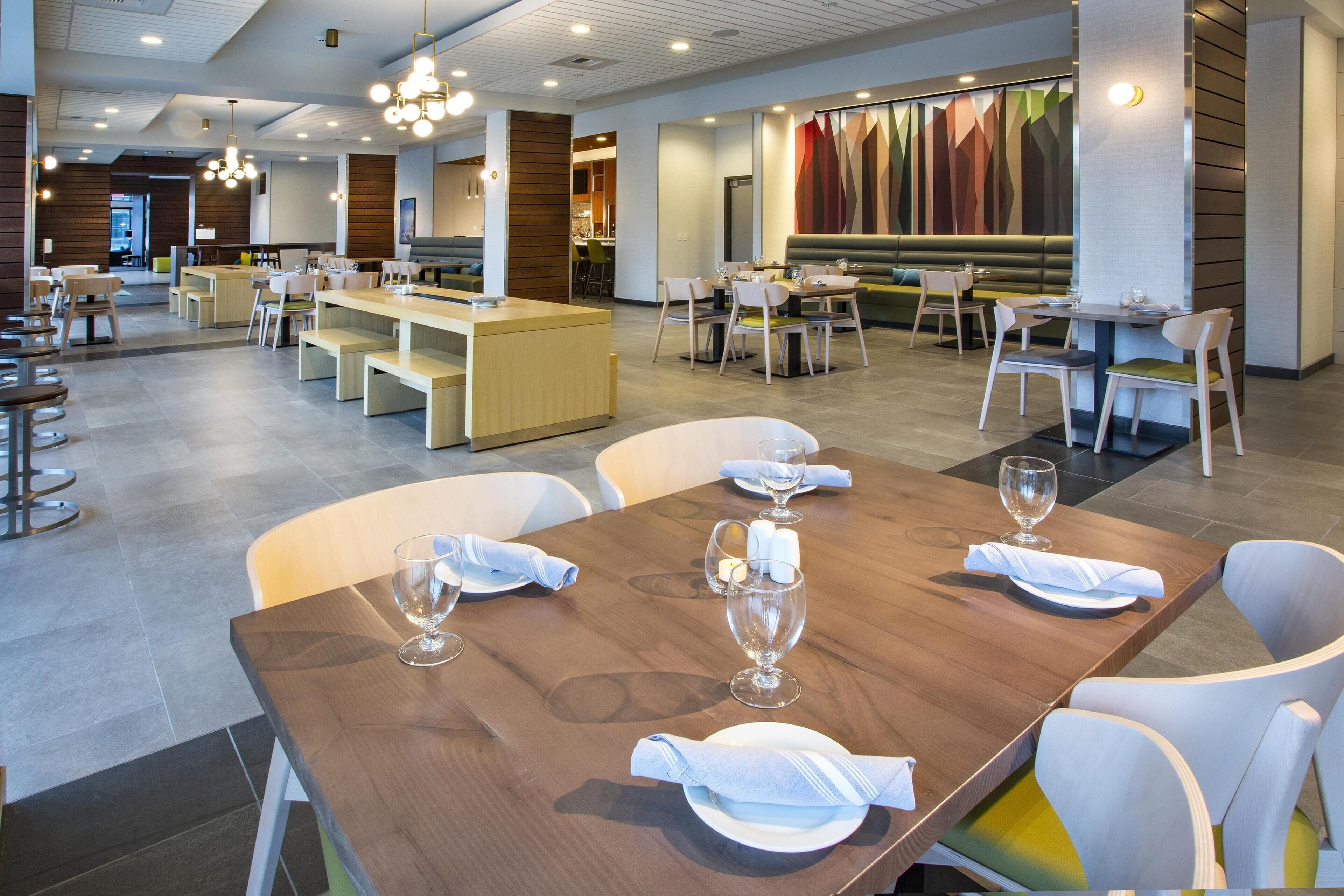 Ein modernes Restaurant mit Holztischen, weißen Stühlen und einem farbenfrohen Wandgemälde.
