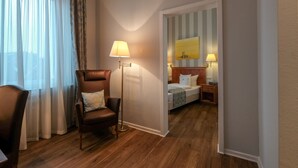 Quarto de hotel com piso de madeira, uma poltrona de couro marrom, um abajur de chão, uma cama com cabeceira listrada e um quadro na parede.