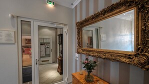 Quarto de hotel com cômoda de madeira, um espelho grande e detalhado e uma porta que dá para um corredor.