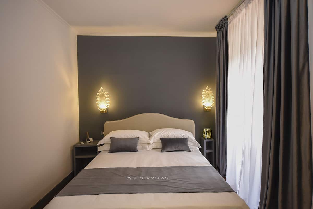 A neatly made bed with a headboard, two bedside tables with lamps, and a dark curtain on the window.
