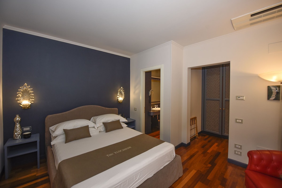 Habitación de hotel con cama, mesitas de noche, un tocador, una silla roja, baño con lavamanos y un armario.