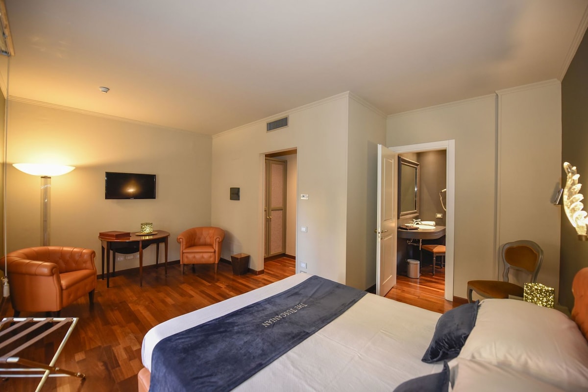 Habitación de hotel con cama, escritorio, silla, televisor y baño visible a través de una puerta abierta.