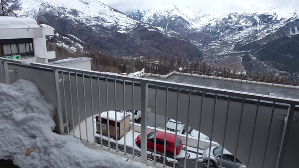 A view from a balcony with a metal railing, overlooking a snowy mountain landscape and a parking area with a red car and a white car.