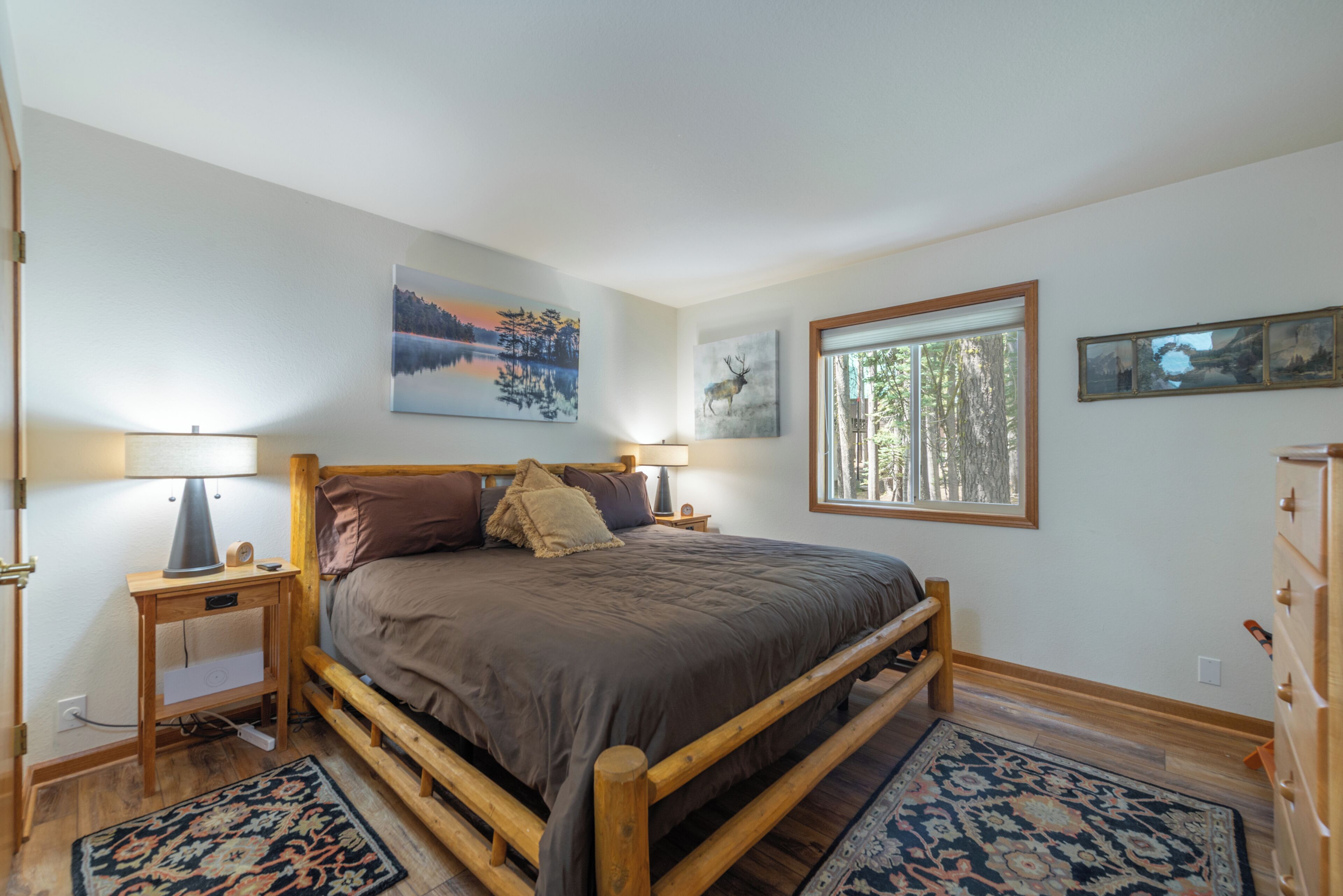 Une chambre à coucher avec un lit en bois, deux tables de chevet, une commode, une fenêtre donnant sur des arbres et un tableau accroché au mur.