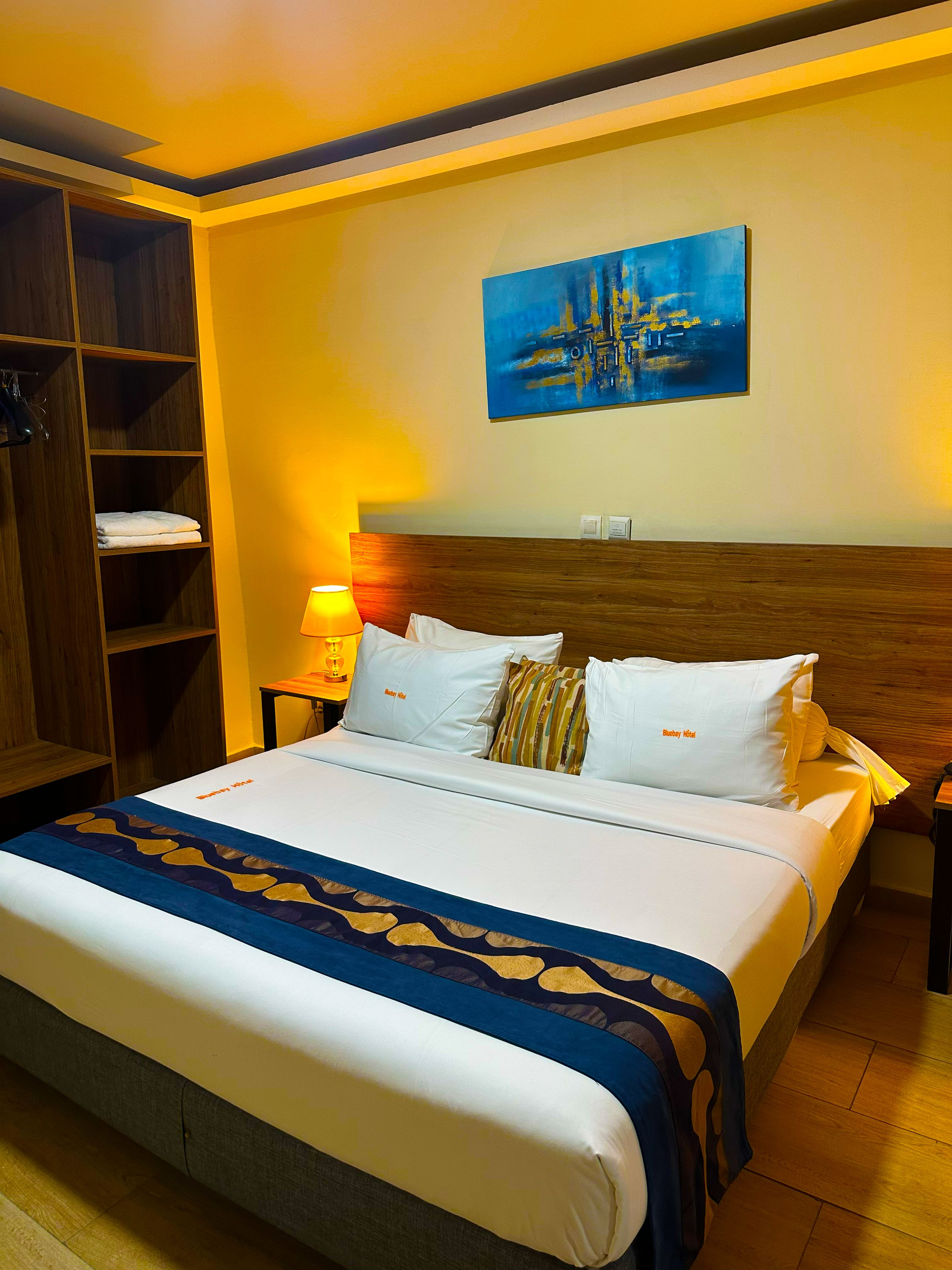 A hotel room with a bed, a nightstand, a lamp, a painting, and a wardrobe.