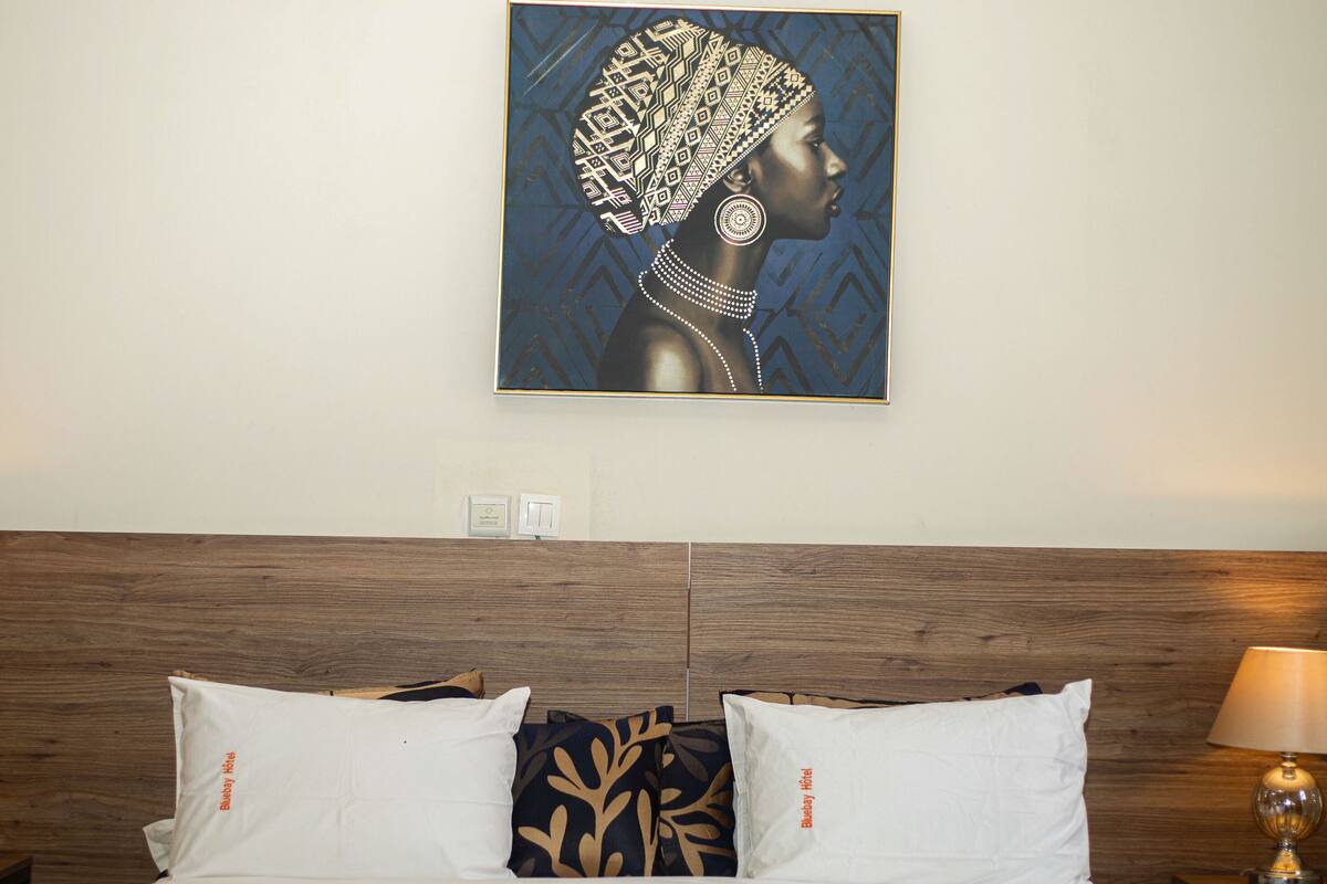 Ein Hotelzimmer mit Holz-Kopfteil, zwei Nachttischlampen, einem Bett mit weißen und gemusterten Kissen und einem gerahmten Kunstwerk an der Wand.