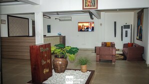 Hall d'albergo con reception, area salotto, piante in vaso e schermo televisivo.