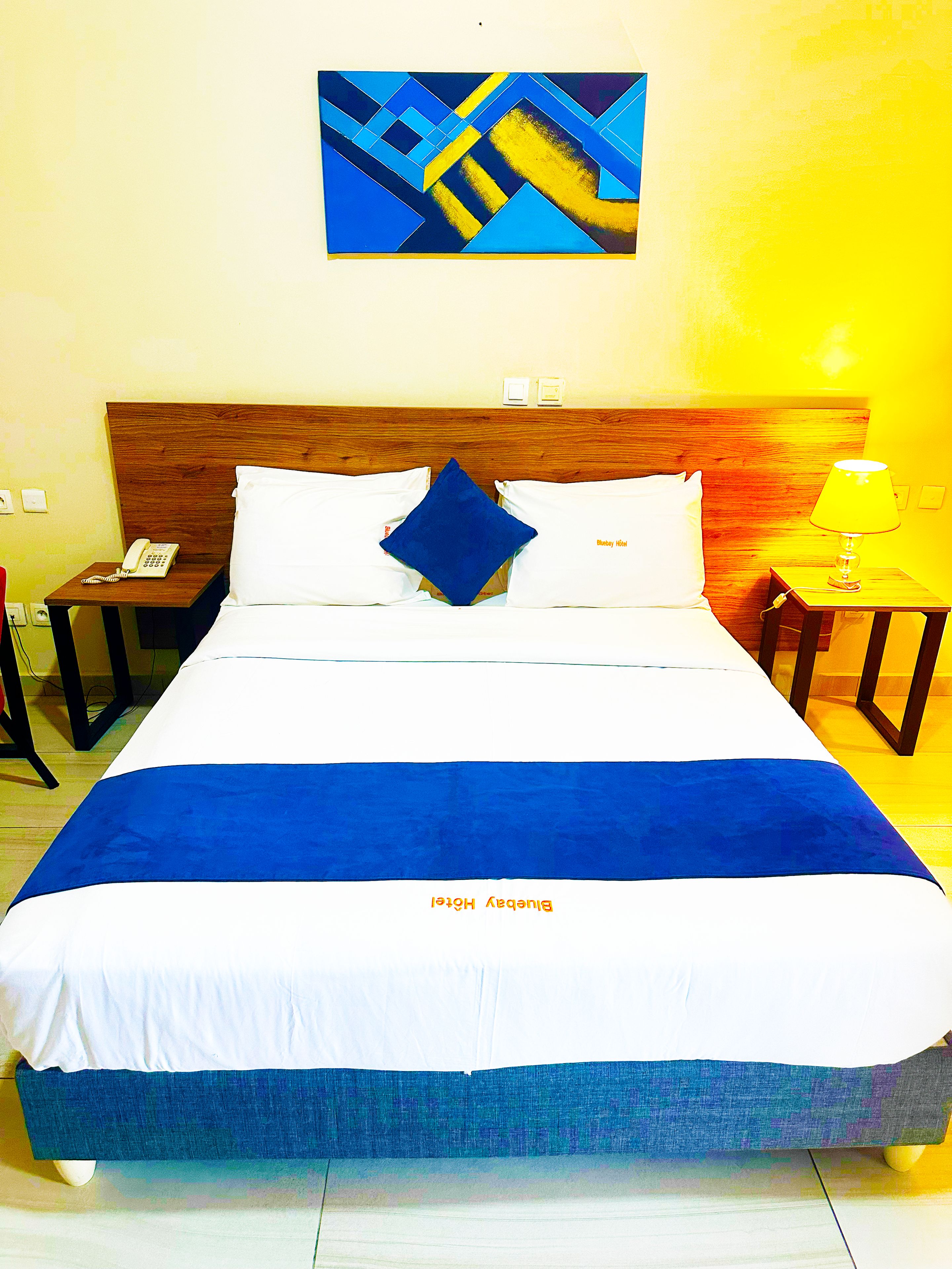 Une chambre d’hôtel avec un grand lit, une tête de lit en bois, une table de chevet avec une lampe, un téléphone et un tableau au mur.