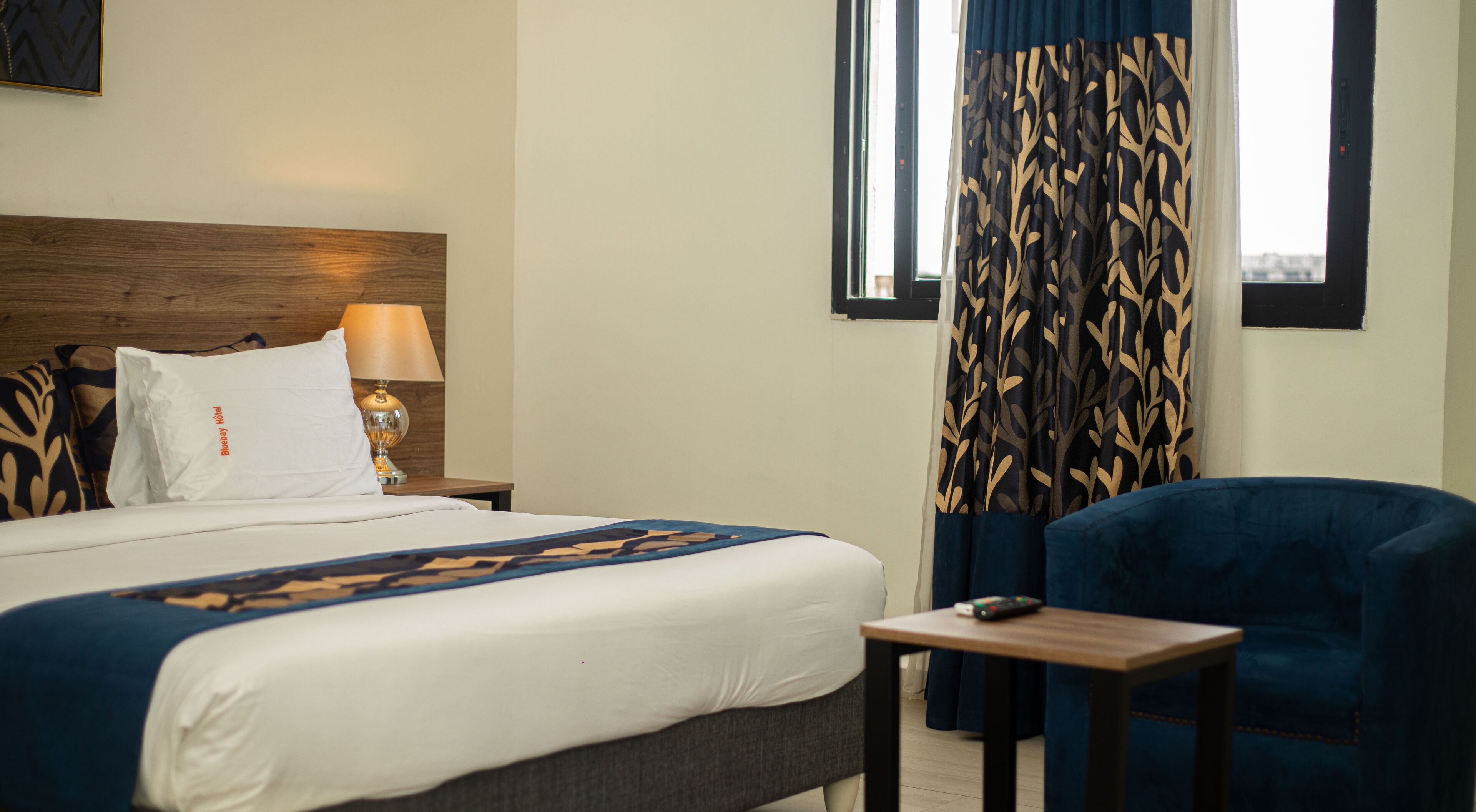Habitación de hotel con cama, mesita de noche con lámpara, silla y ventana con cortinas.