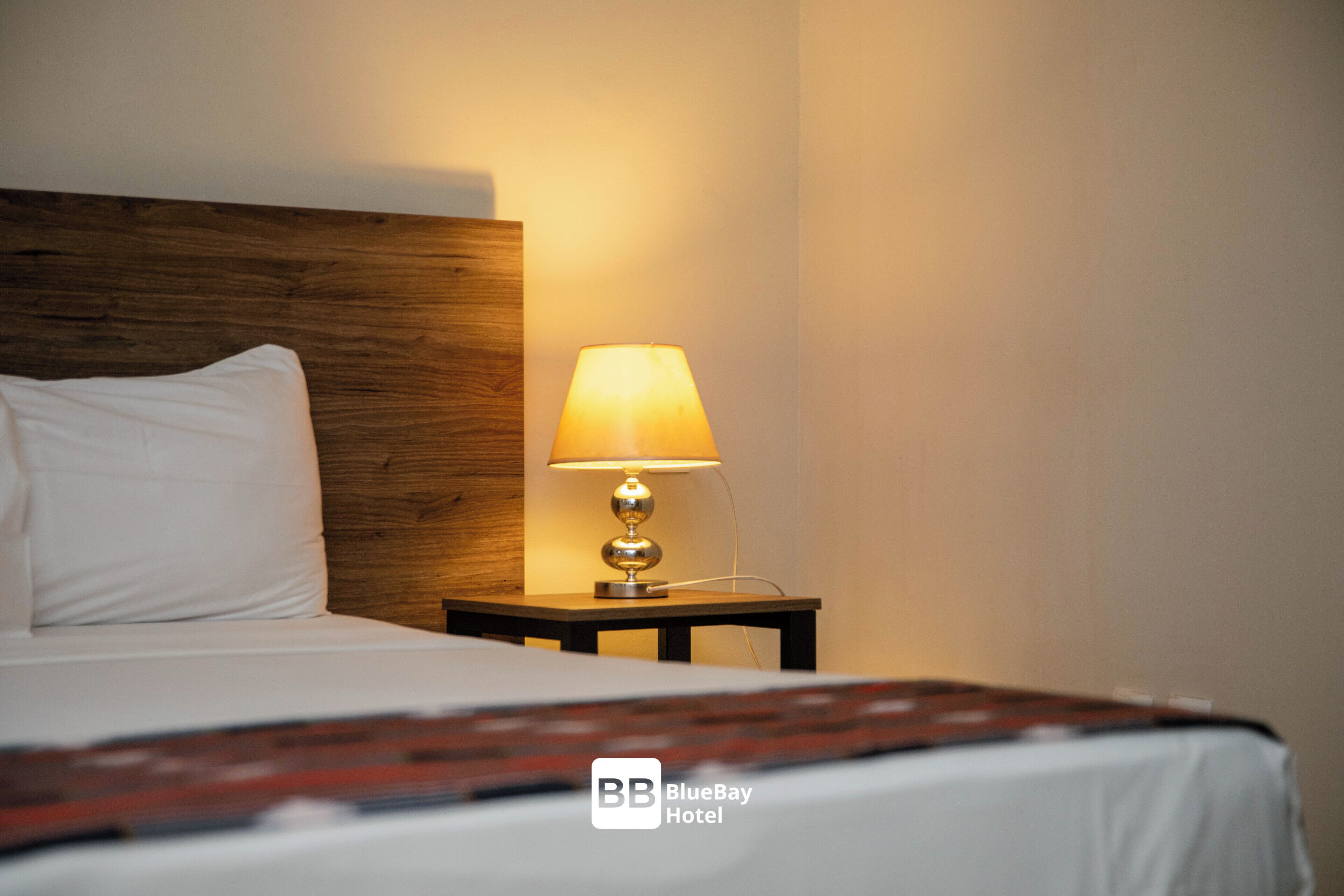 Una habitación de hotel con una cama, cabecera de madera, mesita de noche con una lámpara y una manta en la cama.