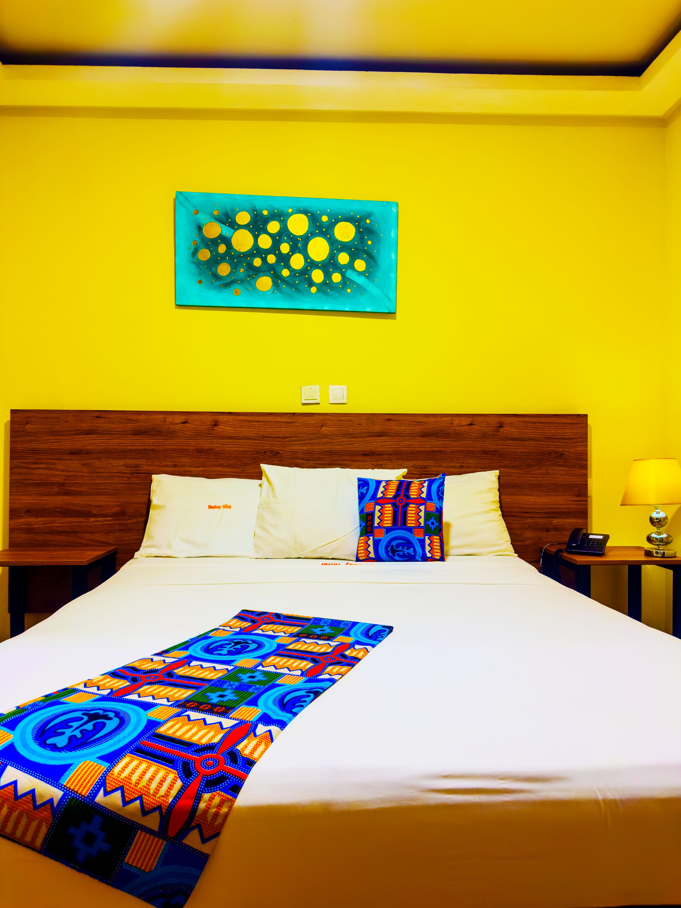 Una habitación de hotel con una cama grande, cabecera de madera, mesita de noche con lámpara y una obra de arte en la pared.