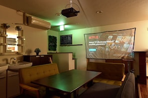 A room with a dining table, chairs, a projector screen displaying a Netflix advertisement, a small plant, and a wall-mounted shelf with a mirror and a sink.