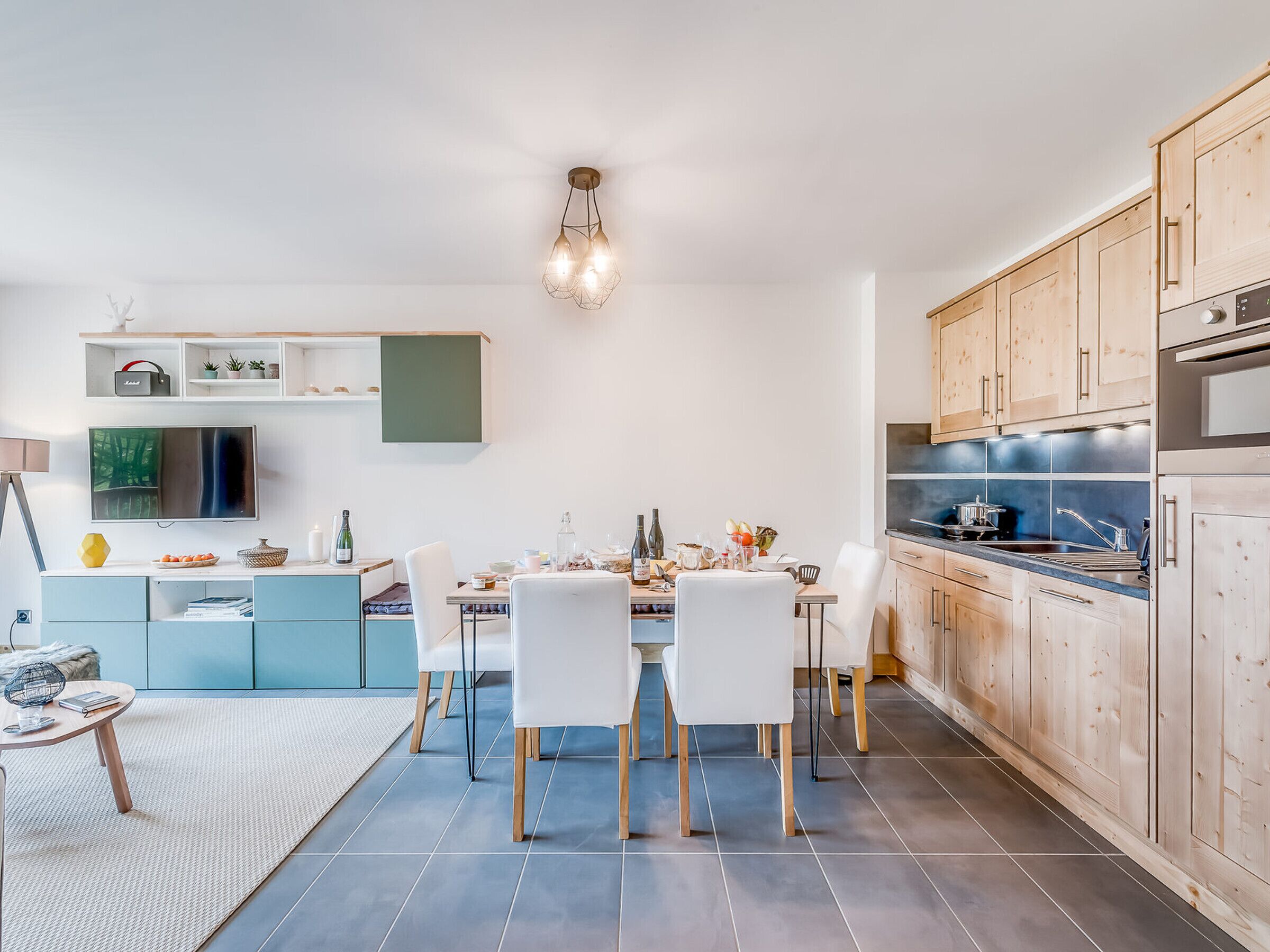 Une cuisine moderne avec des meubles en bois, un coin repas avec des chaises blanches et un téléviseur à écran plat fixé au mur.