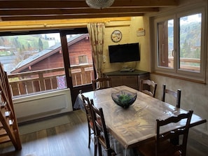 Ein Essbereich mit Tisch, Stühlen, einer Schüssel, einem Fernseher und einem Fenster mit Blick auf ein Holzgebäude und Bäume.