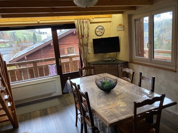 Un comedor con una mesa, sillas, un bol, un televisor y una ventana que ofrece vistas a un edificio de madera y árboles.