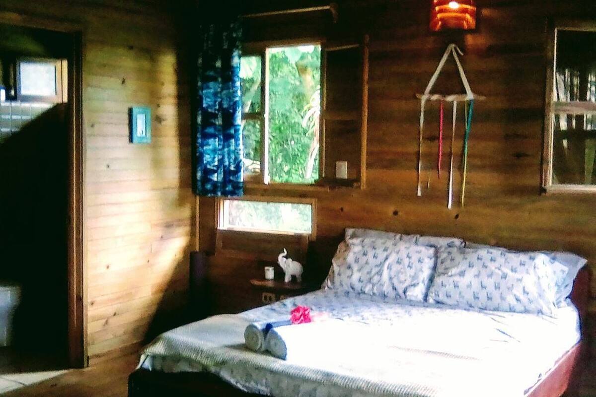 A wooden cabin bedroom with a bed, a window with curtains, a small table with a cup, and a hanging light fixture.