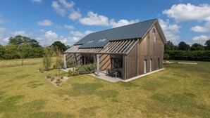 Une maison moderne en bois, dotée d’un toit en pente et de grandes fenêtres, entourée d’un champ verdoyant et d’arbres.