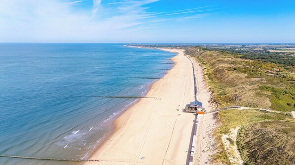 Une vue aérienne d’une plage avec une longue jetée s’étendant dans la mer, une côte sablonneuse et un chemin menant à un bâtiment équipé de panneaux solaires.