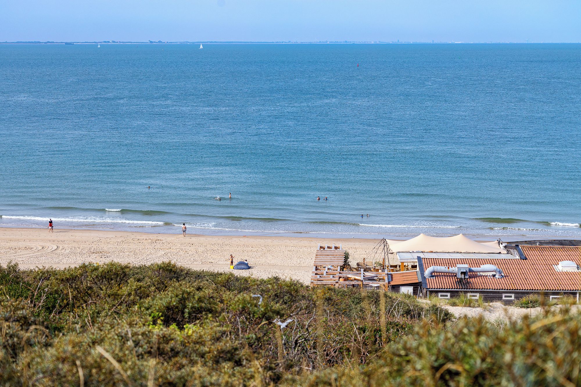 Une plage avec un rivage sablonneux, quelques personnes et un bâtiment avec un toit rouge, visible au premier plan.