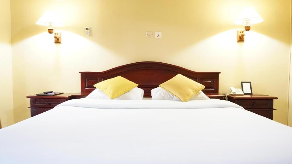 A hotel room with a large bed, two bedside lamps, a nightstand with a phone and a picture frame, and a wall-mounted light switch.