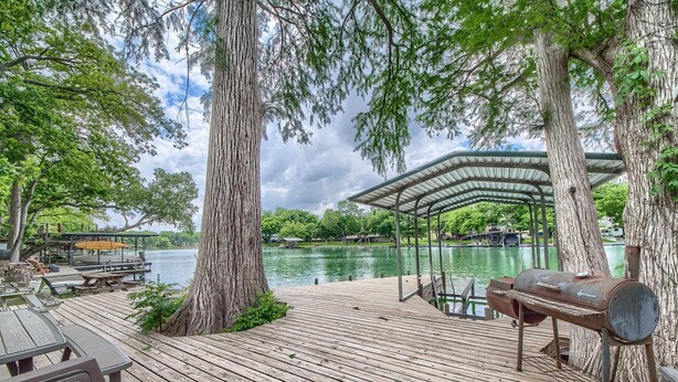 A wooden deck with a barbecue grill, picnic tables, and a covered seating area overlooking a lake.