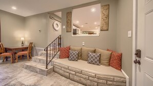 A living room with a stone bench, a dining area with wooden chairs and a table, and a staircase with a railing.