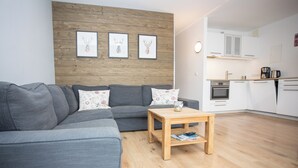 A modern living room with a grey sectional sofa, a wooden coffee table, and a kitchen area with white cabinets and a stove. - Apartement - Am Waltenberg 70-R | Winterberg (Winterberg)