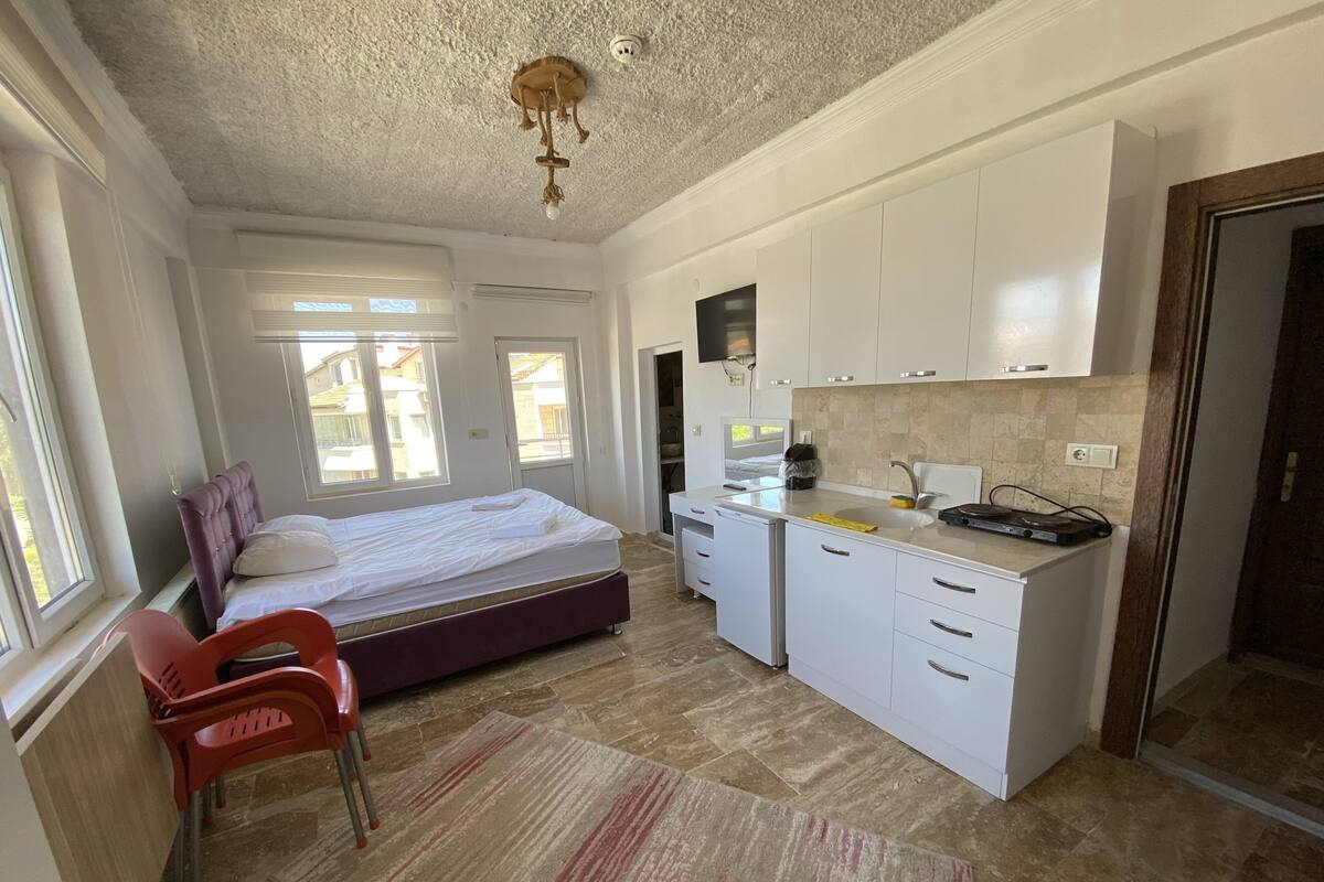 A bedroom with a bed, a desk, and a chair. There is a kitchenette with a sink and a stove. The room has a window and a door.