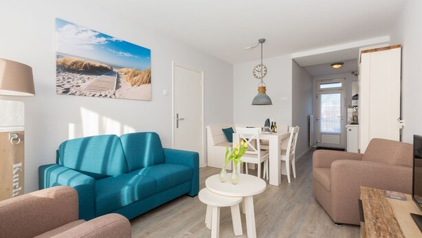 Ett modernt vardagsrum med en blå soffa, en beige soffa, ett vitt matbord med stolar och ett köksområde i bakgrunden.