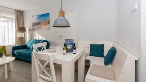 Ett modernt vardagsrum med en turkos soffa, vita fåtöljer och ett vitt matbord. På väggen hänger en tavla, och på bordet står en lampa och en vinflaska.