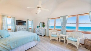 A bedroom with a bed, dresser, TV, and wicker chairs by a large window with a view of the sea.