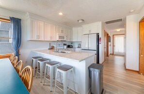 A modern kitchen with white cabinets, a central island with bar stools, a microwave, and a refrigerator. - Wildest Dream - S Nags Head MP 17, Oceanfront, Direct Ocean Access, Air Hockey, Pets OK (South Nags Head)