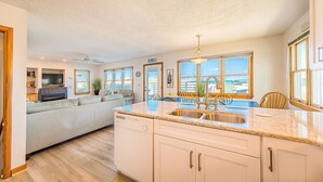 A spacious kitchen with a large island, a dishwasher, and a sink. There is a dining area with a table and chairs, and a view of the ocean through large windows.