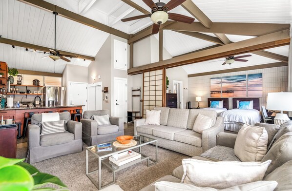 A spacious living area with a high ceiling, exposed beams, and a large window with a sunset view.