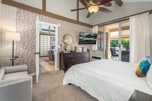 A bedroom with a large bed, a TV mounted on the wall, a ceiling fan, and a view of the outdoors through a sliding glass door.