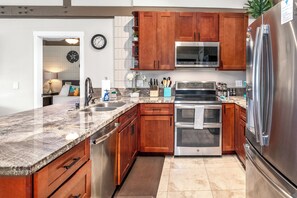 A kitchen with wooden cabinets, stainless steel appliances, a granite countertop, and a clock on the wall.