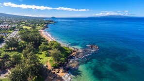 An aerial view of a coastal area with a beach, clear blue waters, and a distant mountain range.