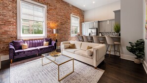 A living room with a brick wall, two sofas, a coffee table, and a kitchen area with cabinets and a refrigerator. - DOWNTOWN 2BR Walk to Superdome,Parade French Quarter (New Orleans)