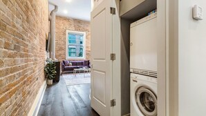 A laundry room with a washing machine, a brick wall, and a sofa in the background. - DOWNTOWN 2BR Walk to Superdome,Parade French Quarter (New Orleans)