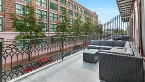 Un balcón con mobiliario exterior, una verja metálica y un edificio con ventanas verdes al fondo.